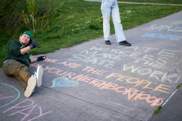 A WFSE member lies back next to their sidewalk chalk art that reads "State workers make the state work! We are the people in your neighborhood." Chalk art of a heart is below.