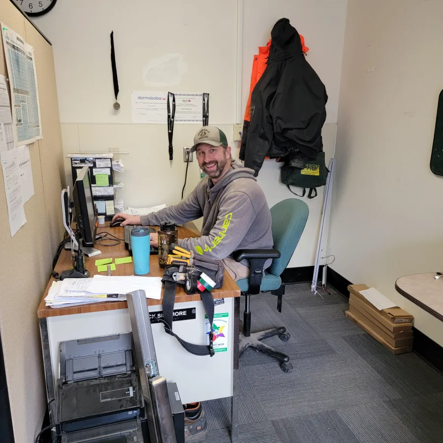A Peninsula College worker sits at a desk next to a tool belt. He is smiling, wearing a grey Carhartt hoodie.