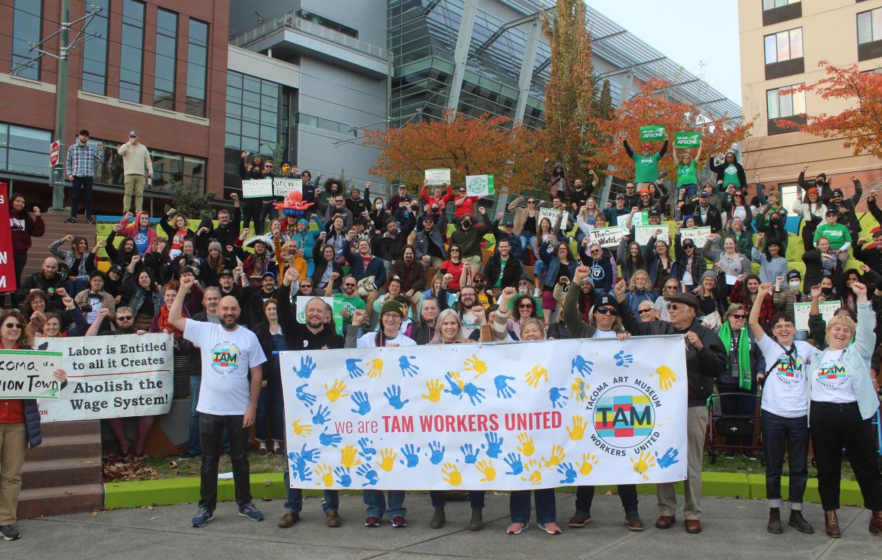 A gathering of hundreds of supporters stand in front of the Tacoma Art Museum, many wearing Tacoma Art Museum Workers United and Washington Federation of State Employees shirts. Many are holding up the solidarity fist. In front, six people hold a TAM sign