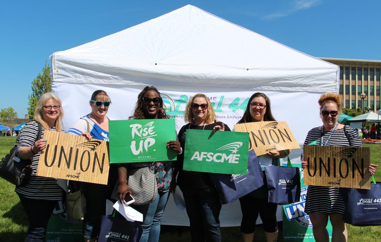 A group of public employees pose with pro-union signs in front of a tent.