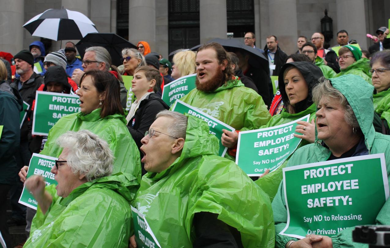 WFSE union members in green rain ponchos hold protest signs about protecting private info during a rally on the Olympia Capitol steps.