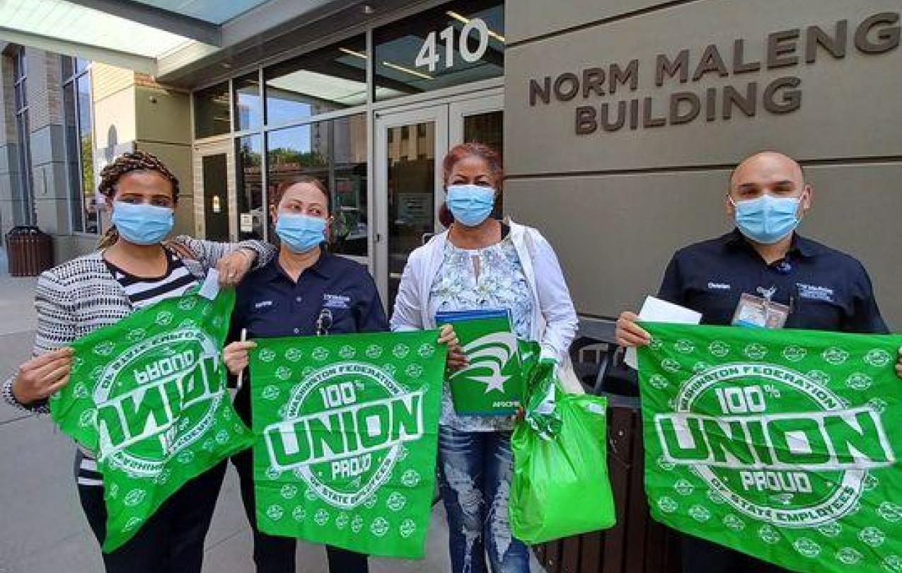 WFSE members & Maleng Cafe employees Ghenet Habtemnichael, Marianna Tereko, Aster Morrow, and Christian Cisneros each hold WFSE green items- three large bandanas and a folder outside the Norm Maleng Building. They are wearing masks.