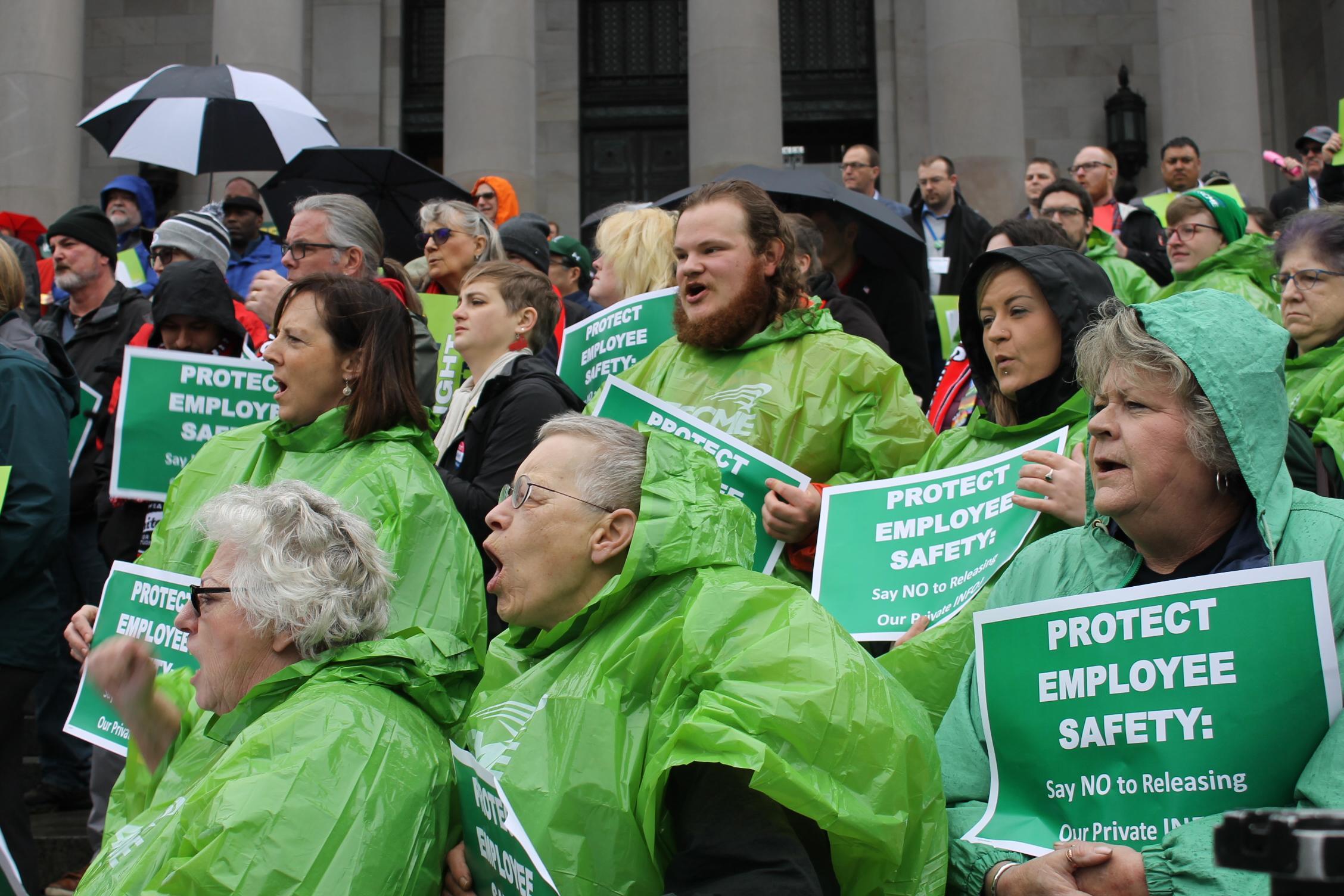 WFSE union members in green rain ponchos hold protest signs about protecting private info during a rally on the Olympia Capitol steps.